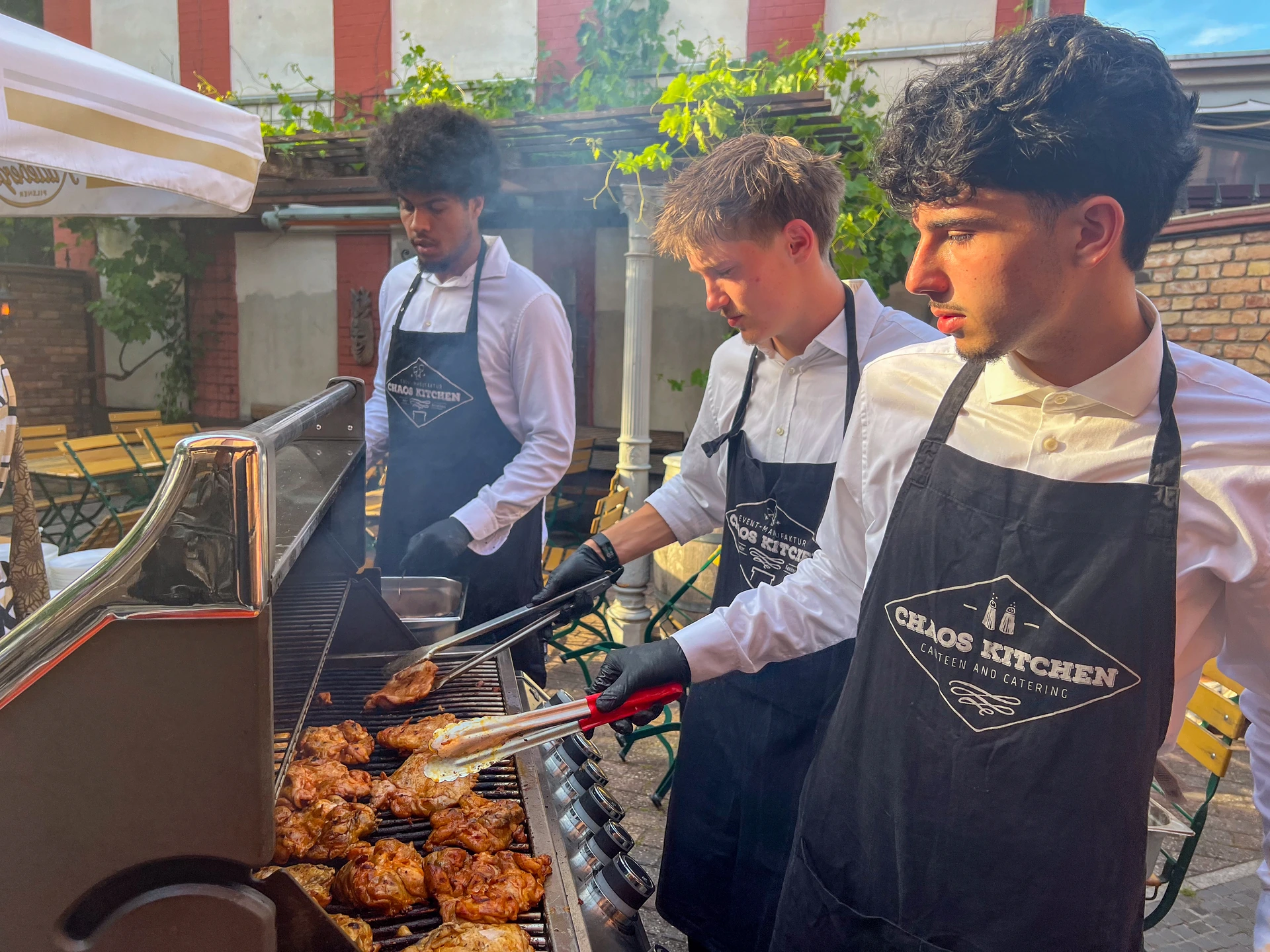 Drei Teammitglieder in weißen Hemden und Chaos-Kitchen-Schürzen bereiten gemeinsam Speisen auf einem großen Außengrill zu.