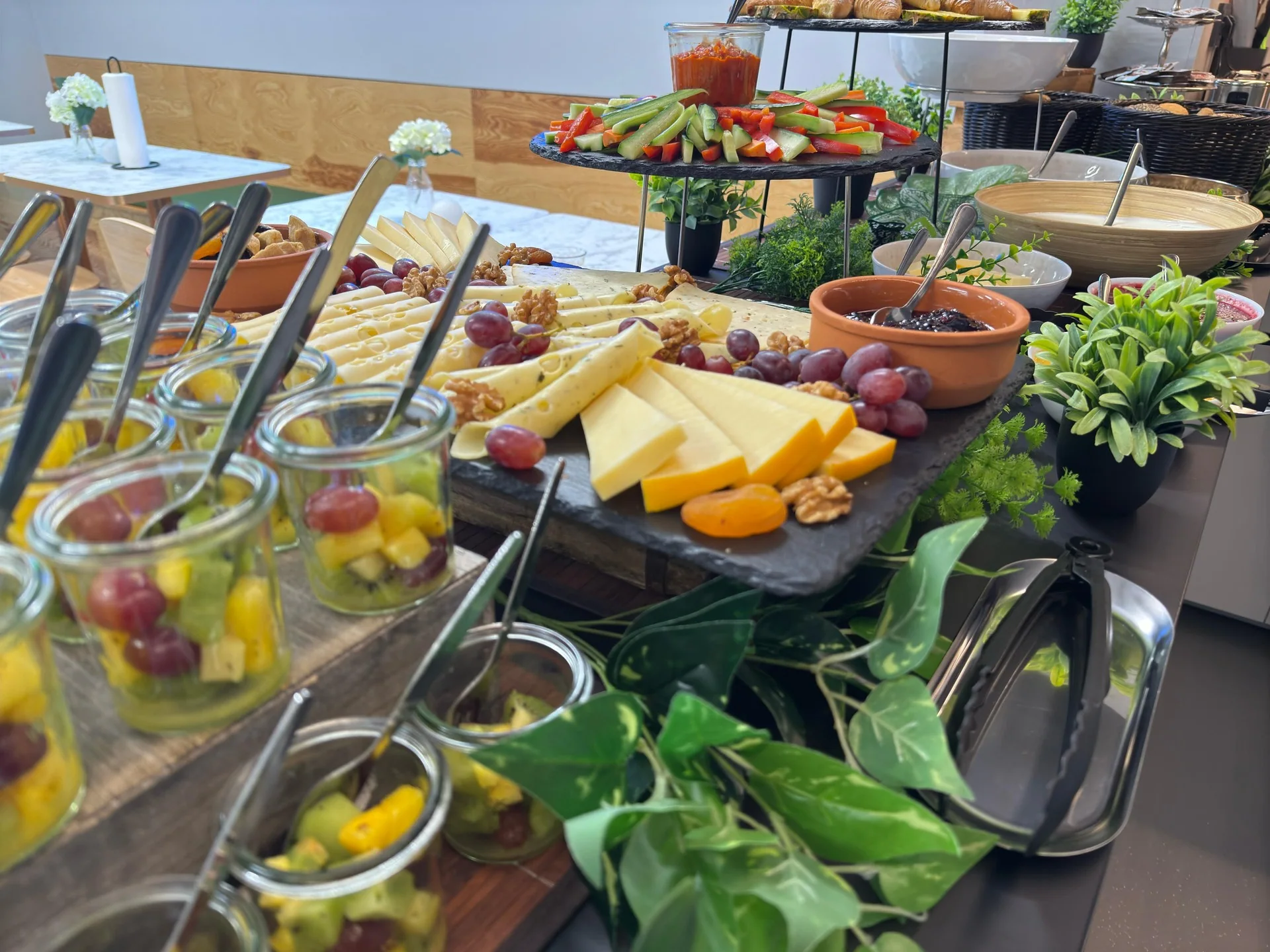 Käseauswahl mit Trauben und Walnüssen neben Obstsalat im Glas; dahinter stehen Gurken- und Paprikasticks auf einer erhöhten Platte.