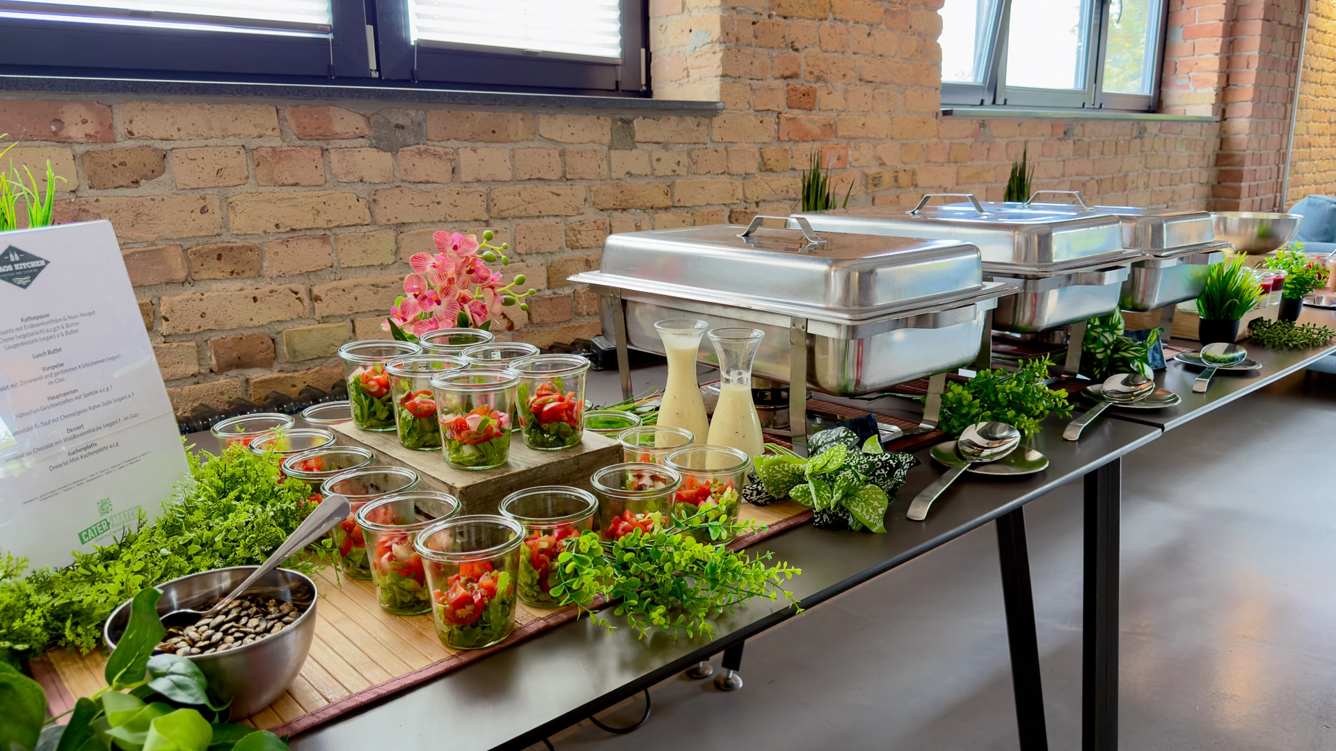 Buffet mit Salaten im Glas, Dressings in Karaffen und Warmhaltebehältern, angerichtet mit Grün und Blumen vor einer Backsteinwand.