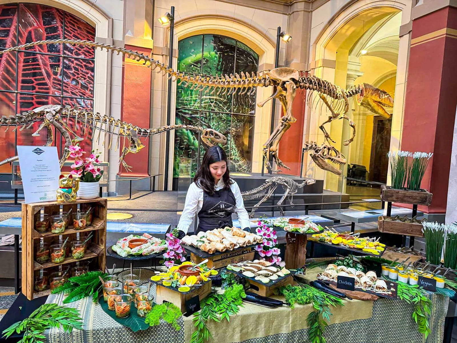 Servicekraft hinter einem mehrstöckigen Fingerfood-Buffet vor Dinosaurierskeletten im Museum für Naturkunde.