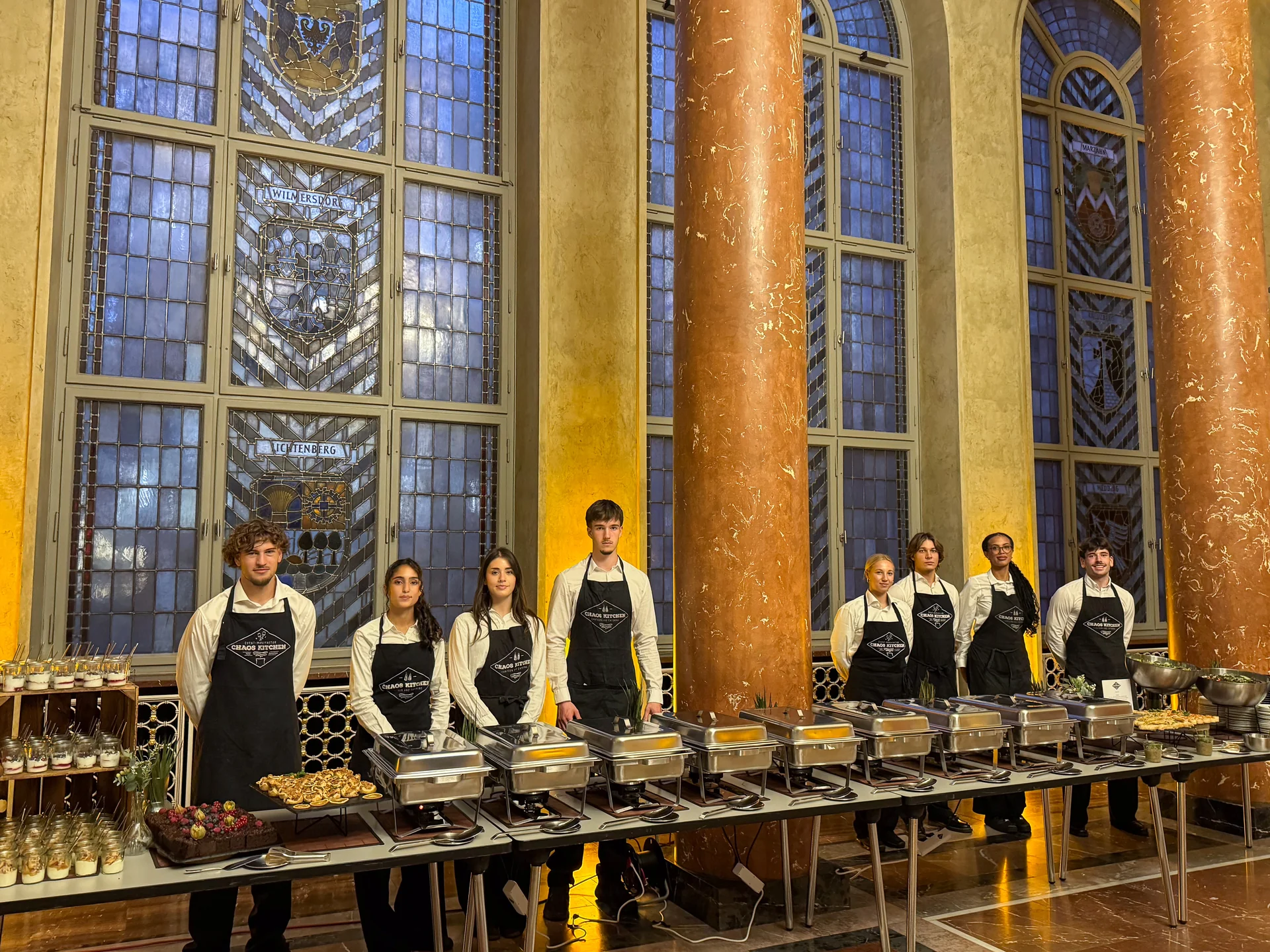 Acht Mitarbeitende von Chaos Kitchen in weißen Hemden und schwarzen Schürzen hinter dem Buffet im Roten Rathaus in Berlin.