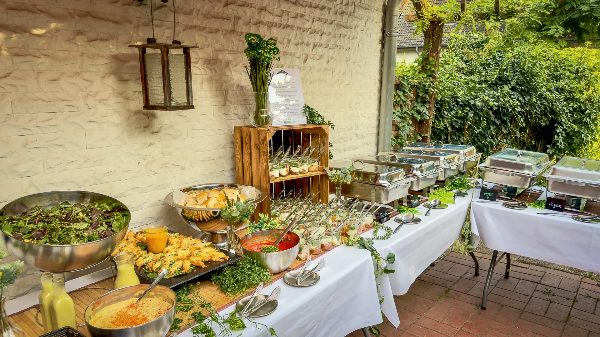 Outdoor-Buffet mit Salatschüssel, Brot, kleinen Speisen im Glas und Warmhaltebehältern auf weiß gedeckten Tischen.