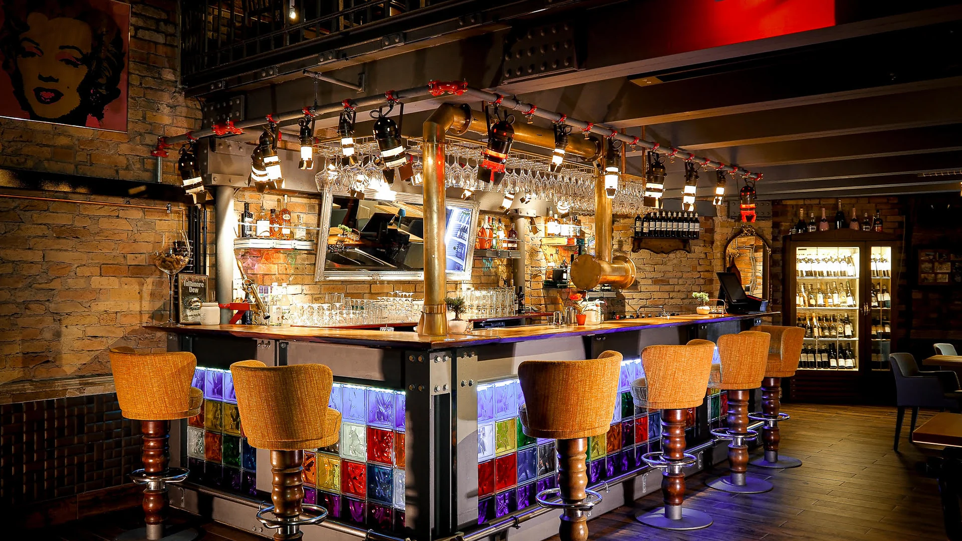 Bar im XELOR Kesselhaus mit Backsteinwänden, Kupferrohren, hängenden Gläsern und beleuchteten Glasbausteinen.