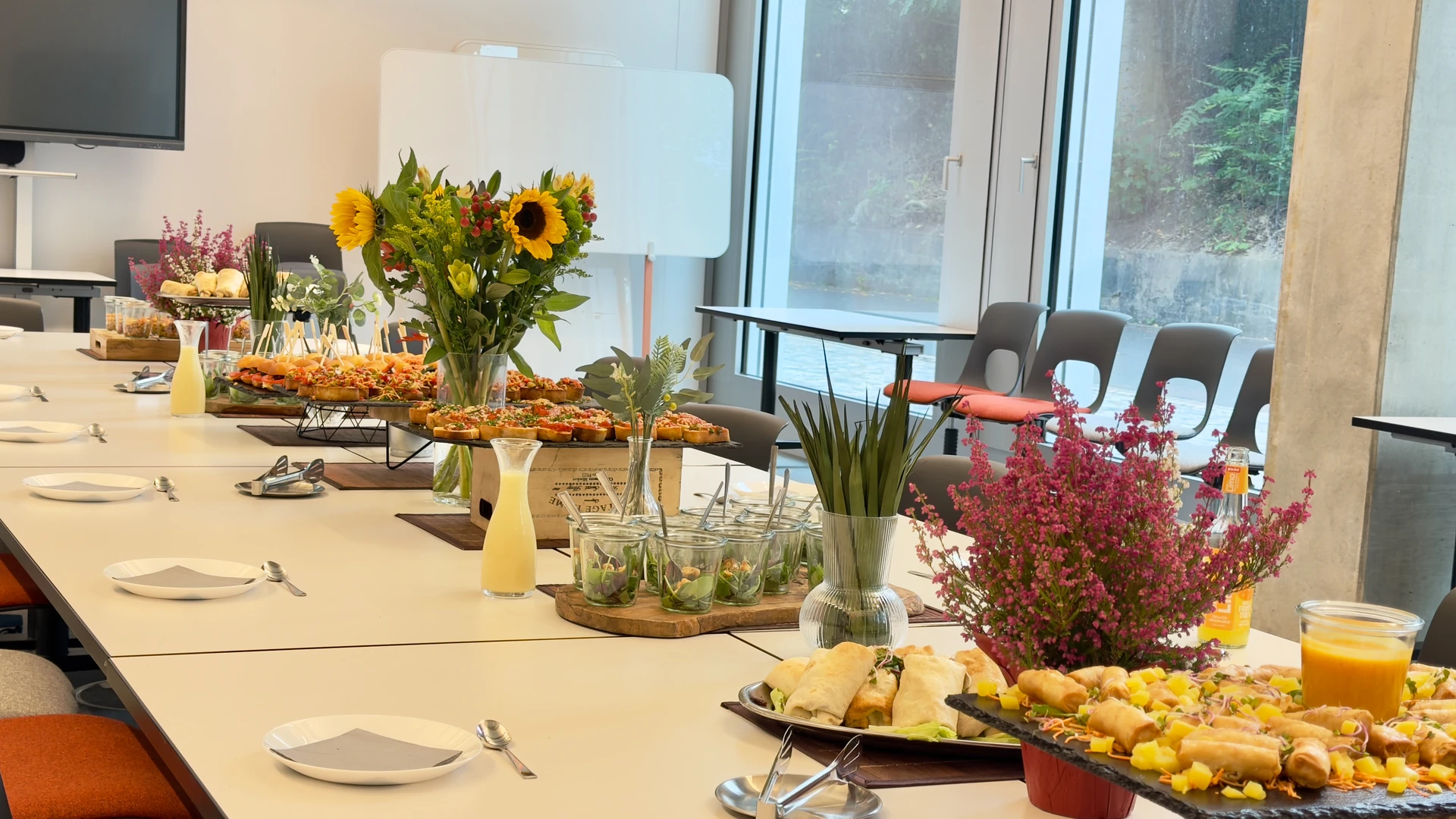 Gedeckter Besprechungstisch mit Fingerfood auf erhöhten Platten, Salaten im Glas, Getränkekaraffen und Blumen.