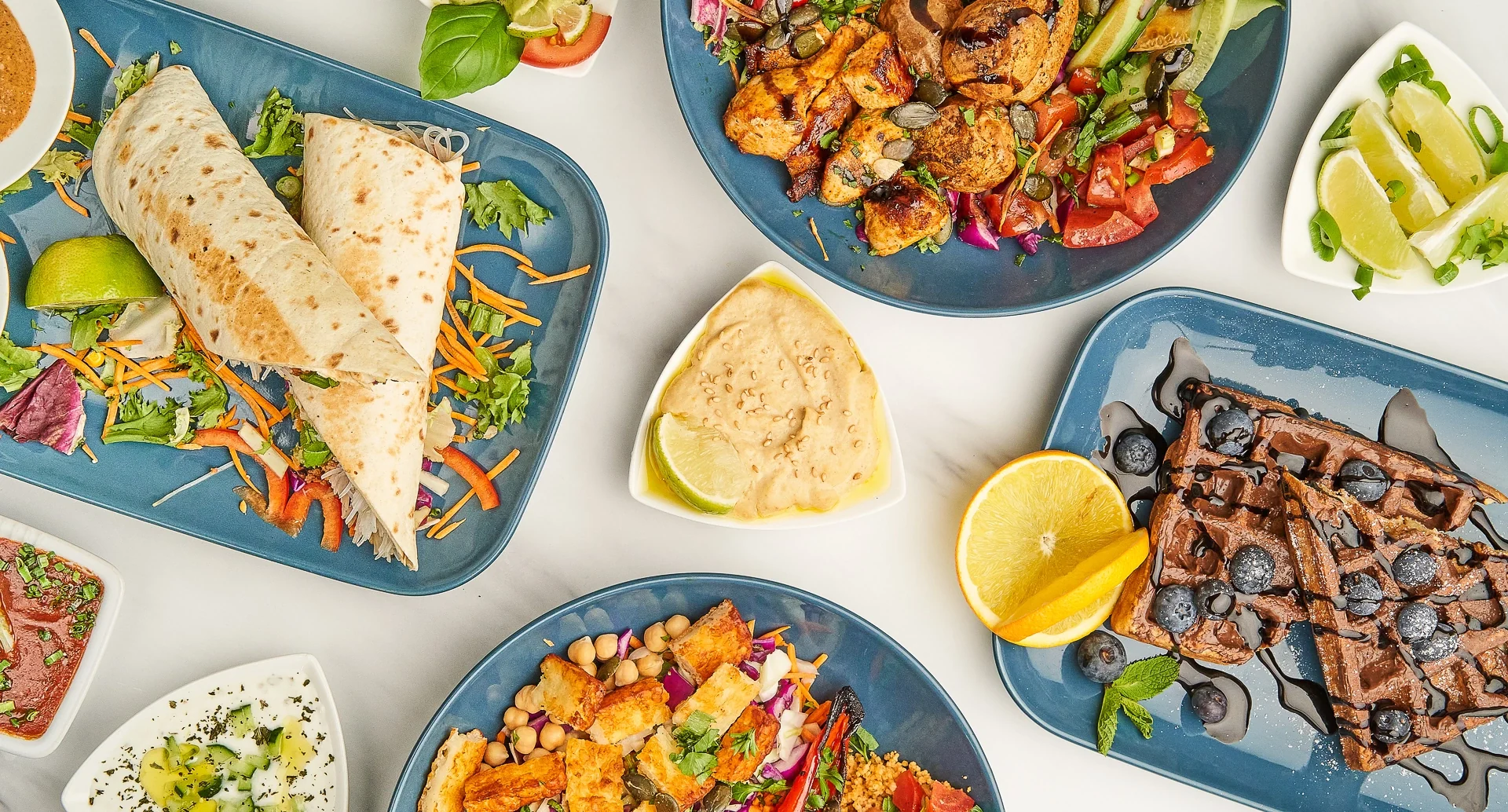 Wraps, bunte Tellergerichte, Waffeln und Dips auf blauen Tellern, von oben fotografiert.