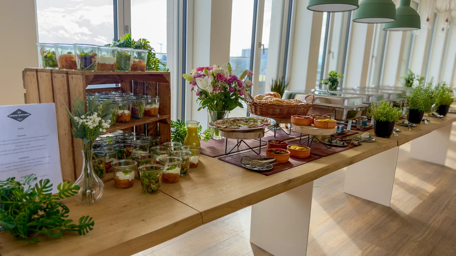Business-Buffet mit kleinen Gläsern auf Holzkisten, Blumen, Brot und Warmhaltebehältern vor großen Bürofenstern.