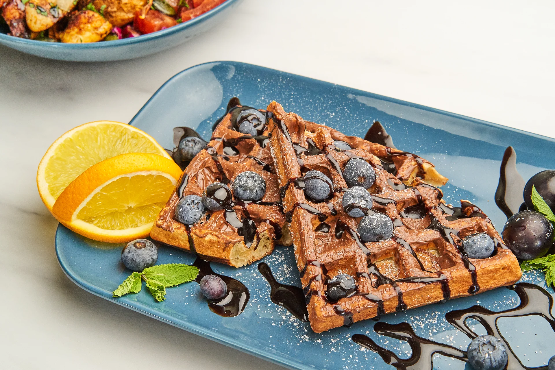 Waffelstücke mit dunkler Sauce, Beeren und Orangenscheiben auf einem blauen Teller.