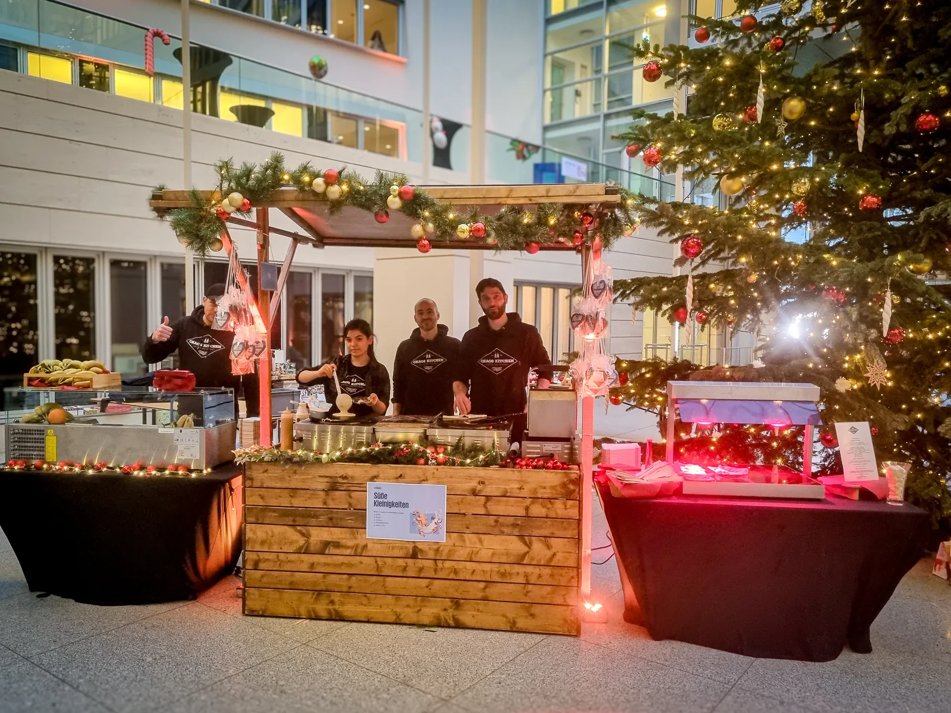 Vier Teammitglieder an einem weihnachtlich geschmückten Crêpe-Stand neben einem beleuchteten Tannenbaum.