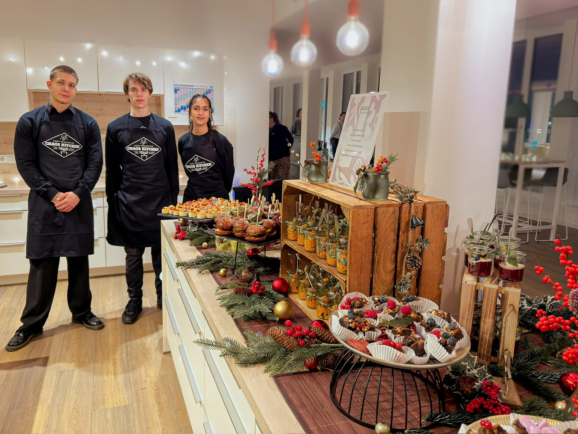 Drei Personen in Chaos-Kitchen-Schürzen neben einem weihnachtlich dekorierten Fingerfood-Buffet.
